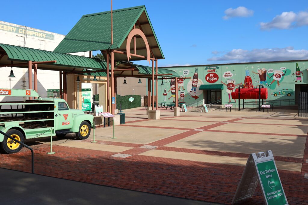 Open, outdoor courtyard containing a green soda delivery truck and a large Dr Pepper painted mural of anthropomorphic bottles and cans. 
